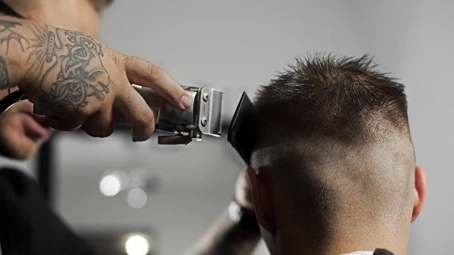 Tattoed barber makes haircut for customer at the barber shop by using hairclipper, man's haircut and shaving at the hairdresser, barber shop