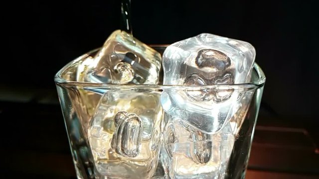 barman pouring whiskey in the glass with ice cubes on wood table and black dark background, focus on ice cubes, whisky relax time on warm atmosphere