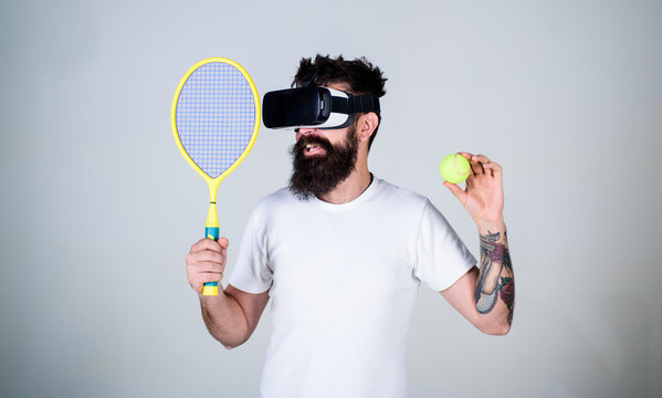 Guy with VR glasses play tennis with racket and ball. Man with beard in VR glasses play tennis, grey background. Virtual sport concept. Hipster on smiling face use modern technology for sport games