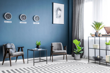 Two grey armchairs, metal rack with decor, window with drapes and fresh plants placed in dark living room interior - obrazy, fototapety, plakaty
