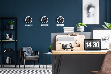 Motorcycle model placed on study corner desk with laptop and fly poster in dark industrial flat interior with three clocks on the wall, metal rack and grey armchair