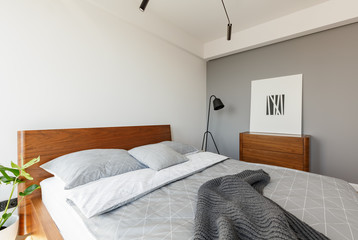 Grey blanket and pillows on wooden bed in modern hotel bedroom interior with poster. Real photo