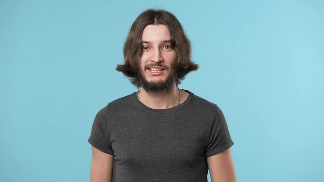 Portrait of cheerful unshaved man 20s wearing casual gray t-shirt and necklace making faces and having fun, over blue background. Concept of emotions