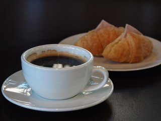 Black coffee in white glass is placed in the front of the Croissant on a wooden table in a coffee shop.