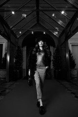 Gorgeous young woman with long brunette hair dressed in gray jumper, trousers and luxurious fur coat, holding handbag, posing on red carpet against building entrance and potted trees on background.
