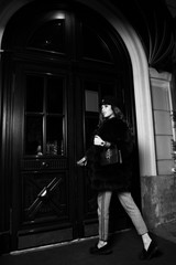 Fototapeta na wymiar Glamorous woman with long wavy brunette hair dressed in gray trousers and luxury fur coat, holding elegant handbag, opening antique wooden door to enter building. Gorgeous female model in outerwear.