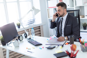 A man is sitting in the office at the table, talking on the phone and flipping through documents.