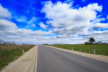 Asphalt highway in countryside with gravel shoulders on both sides and cloudy blue sky as background