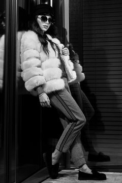 Full body portrait of fashionable young long haired brunette woman wearing large round sunglasses, cap, luxurious white fur coat, standing with her back leaning on large glass window and posing.