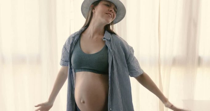 Excited woman in expectancy wearing top and hat and dancing emotionally having fun at home alone