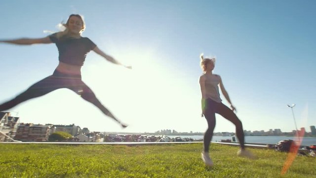Two young slim women performing synchronous jumping on the grass under the sun