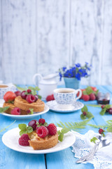 Delicious summer breakfast, homemade pastries and fragrant coffee. On a blue wooden background, berries and green leaves and flowers. Copy space.