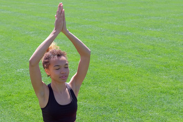 A beautiful young African-American woman in a black T-shirt closed her eyes and raised her hands up against the background of the green grass. Modern sports lifestyle. Copy space.