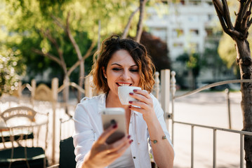 Fototapeta premium Portrait of a smiling young woman drinking coffee and using smartphone outdoors.