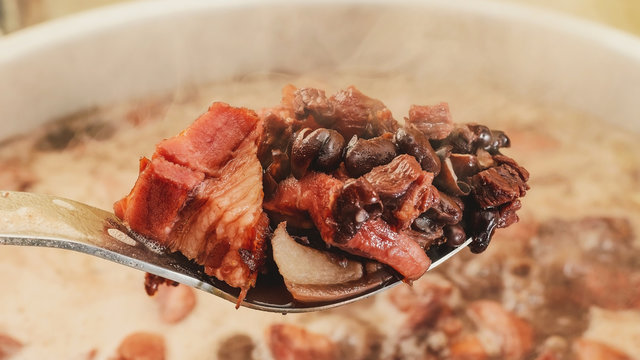 Spoon taking out of the pan ingredients of Feijoada, Brazilian cuisine. Spoon with black beans, pork meat and bacon. Feijoada cooking in the pan on the background with steam around.