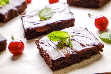 Square pieces of homemade brownies with dark chocolate spread paste, cherry and mint leaves on white table top. High calorie pastry. Cocoa based sweets, bad for figure. Close up, background, top view.