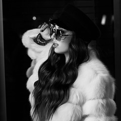 Waist up portrait of gorgeous female model wearing large round sunglasses, cap and stylish white half-length fur coat posing on street. Lovely smiling long haired woman in fashionable outfit.