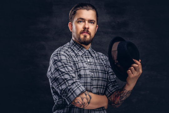 Portrait of a tattoed middle age hipster man with beard and hairstyle dressed in a checkered shirt pose with a hat in hand.