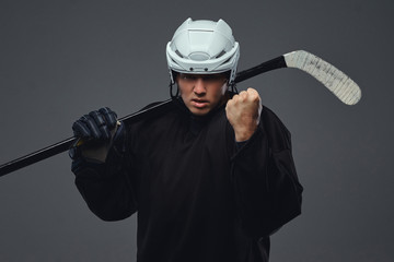 Angry professional hockey player in black sportswear standing with a hockey stick on a gray...