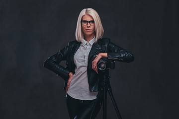 Smart blonde female photographer in trendy clothes posing while leaning on a professional camera with a tripod at a studio, looks at the camera.