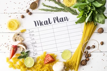 Tuinposter A meal plan for a week on a white table among products for cooking - pastas, basil, vegetables, lime, seeds, nuts and spices. Top view, flat lay, copyspace © svitlini