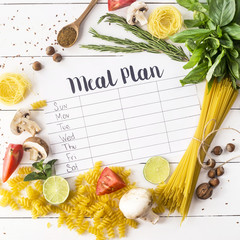 A meal plan for a week on a white table among products for cooking - pastas, basil, vegetables,...