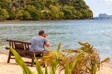 A man sitting on a bench by the sea takes pictures of the seascape.