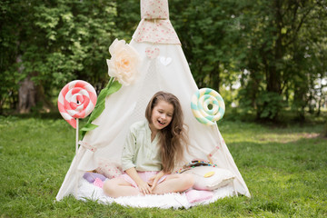 Little girl lying and playing in a tent, children's house wigwam in park. Happy mothers day. Child with big donuts and heart-shaped pillows. Summer, childhood concept. © MartaKlos