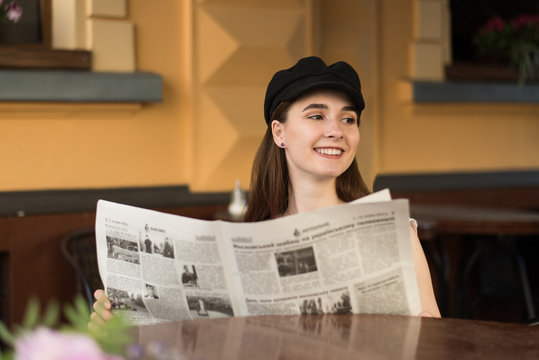 Young woman having a breakfast reading newspaper outdoors at the typical french cafe terrace in France. Playful girl with perfect long hairs posing outdoor. Wearing wool cap. Street fashion look. 