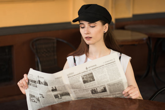 Young woman having a breakfast reading newspaper outdoors at the typical french cafe terrace in France. Playful girl with perfect long hairs posing outdoor. Wearing wool cap. Street fashion look. 