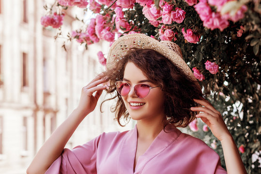 Outdoor close up portrait of young beautiful happy smiling curly girl wearing stylish pink sunglasses, straw hat, dress. Model posing in street near blooming roses. Summer fashion concept