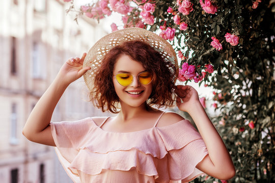 Outdoor close up portrait of young beautiful happy smiling curly girl wearing stylish yellow sunglasses, straw hat, pink blouse with ruffles. Model posing near blooming roses. Summer fashion concept