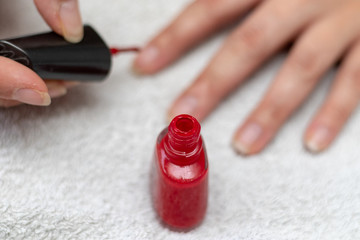 Small bottle of red nail enamel in front of the hands of a woman coating the nails with red lacquer