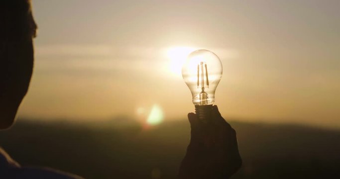 A woman holds a house light bulb in which the sun is setting inside. Concept of: renewable energy, nature, electricity, love for the environment and for the planet.