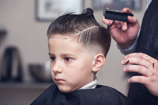 Barber making hairstyle to a Caucasian boy using scissors and hairbrush.
