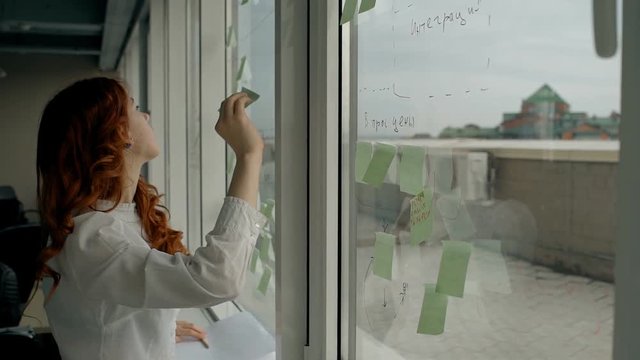 Beautiful young woman glues stickers on window standing in modern office, female employee in white shirt works with sticky notice during working day in bright room. Concept: professional, modern