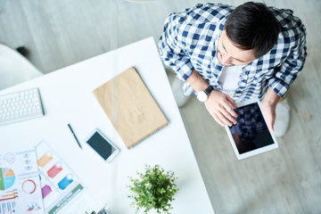 From above shot of casual man holding tablet and standing in modern creative office near table. 