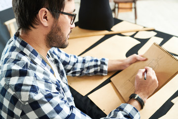 Side view of casual man in glasses creating design for dress in notepad sitting at desk in studio. 