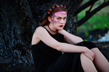 A young beautiful woman with a violet shine on her face sits near a burnt tree