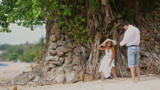 Man takes photo of beautiful woman in hat and white dress on a camera Outdoors Under the Tree with roots. Happy smiling man and woman on beach. slow motion, 3840x2160