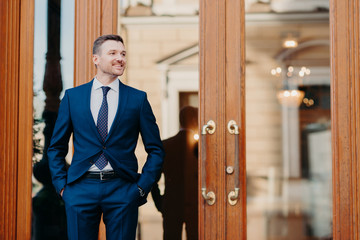 Good looking satisfied business worker wears elegant luxury suit, stands near entrance to...