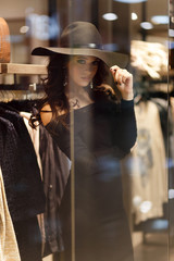 Fototapeta na wymiar Young beautiful girl with perfect make-up, wearing a black hat and elegant black dress, posing near glass storefront of the shopping center.