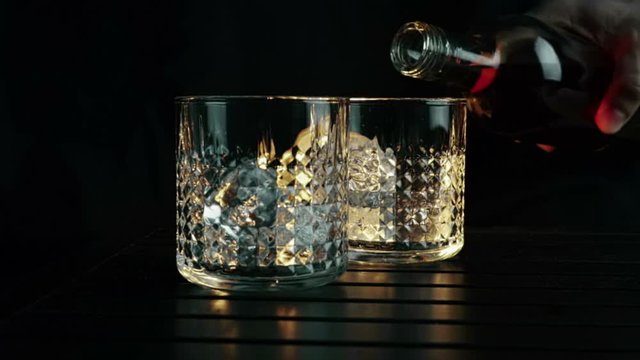 barman pouring whiskey in the two glasses with ice cubes on wood table and black dark background, focus on ice cubes, whisky relax time on warm atmosphere