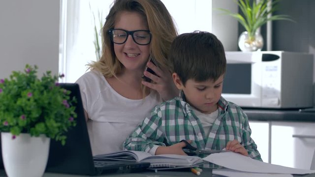 woman combine upbringing child and work, mother with computer talk on mobile phone while son plays beside her at kitchen