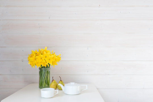 Yellow daffodils in glass jar on white table with two green pears and white teapot against rustic white wood panelled wall