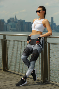 Beautiful young fitness model wearing stylish sportswear posing on the pier with city on the background.