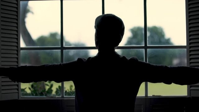 The silhouette of a woman who opens the shutters of a window to let light into a dark room in her home.