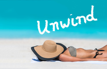 UNWIND word written on sky above beach travel bikini suntan woman sleeping relaxing covering face with hat doing siesta. UNWIND text in blue ocean copyspace above. Summer and sun vacation holidays.