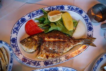 Fresh grilled whole sea bream fish with roasted potato, green salad, tomato and yellow lime on white oval plate with blue pattern at Galata bridge