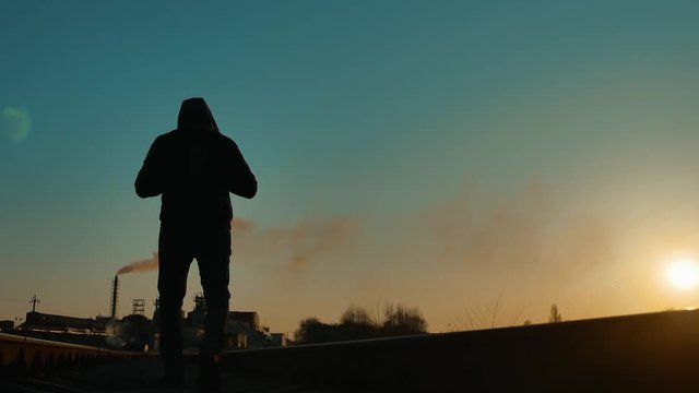 man hiker homeless person traveler walking by railroad silhouette road. man with a backpack going to sunset silhouette against a background of an industrial zone plant pipe smoke. man traveler concept